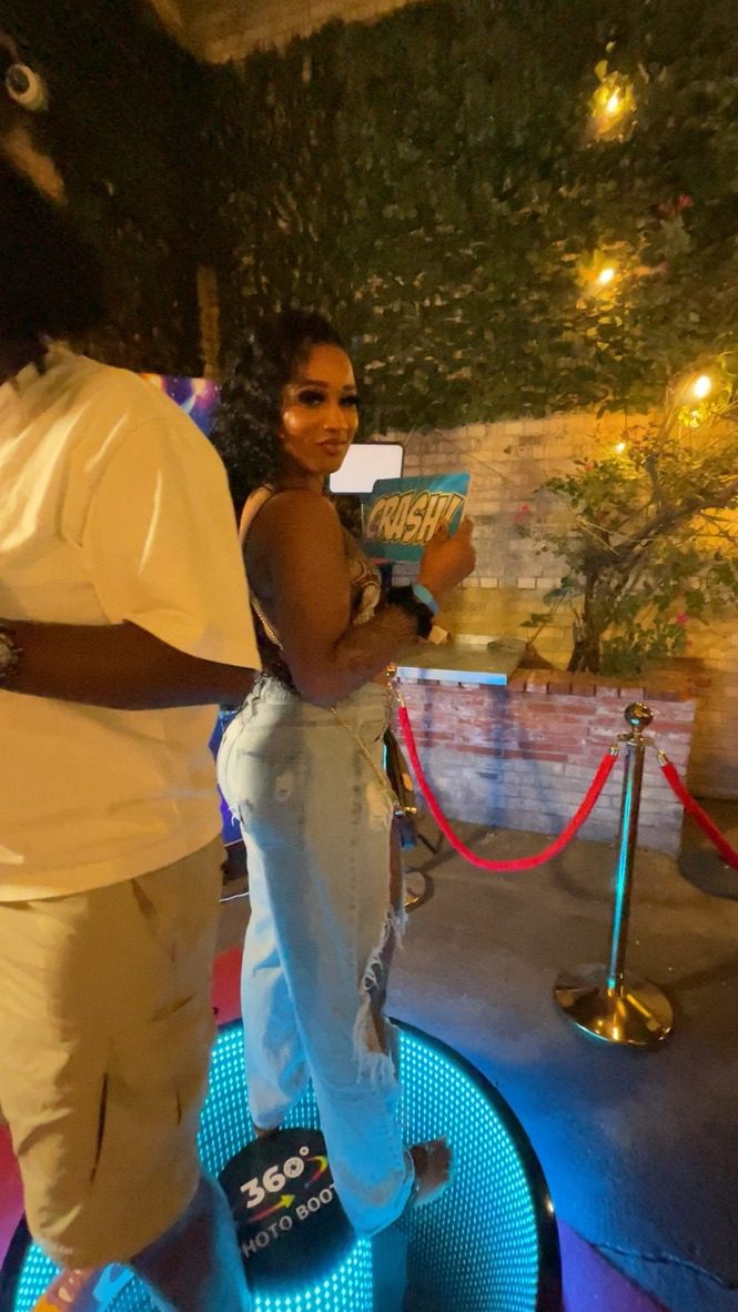 Guest standing on a glowing 360 photo booth platform at an outdoor evening event, holding a party sign while recording a video.