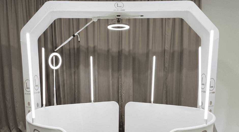 Modern white 360-degree photo booth setup with an overhead frame, mounted ring light camera arm, vertical LED light tubes, and curved panels in front of a neutral curtain backdrop.
